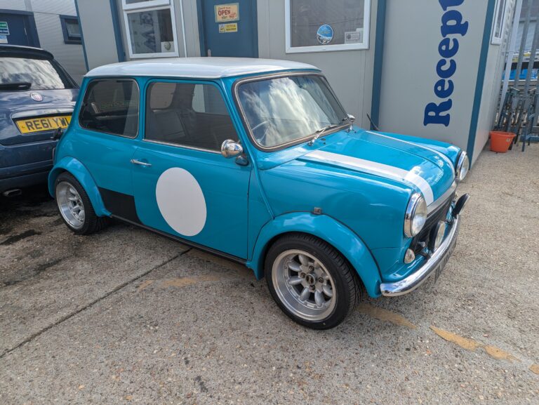 Bright blue classic Mini with white racing stripes, white roof and alloy wheels outside Trickett Welding.