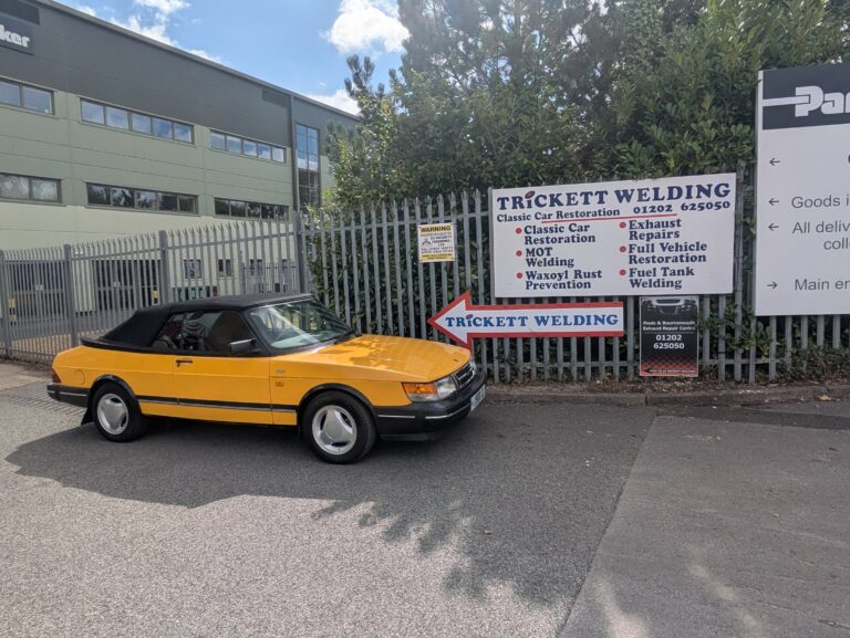 Yellow classic Saab 900 convertible with black soft top outside Trickett Welding in Poole.
