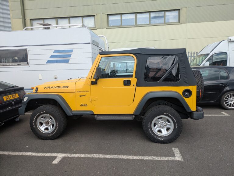 Yellow Jeep Wrangler Sport 4x4 with black soft top and off-road tyres parked outside the workshop.